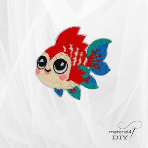 Könnte beinhalten: Gestickter Aufnäher mit einem Cartoon-Goldfisch mit großen schwarzen Augen, roten Flossen und blauen und türkisfarbenen Akzenten. Der Aufnäher hat ein beiges Gesicht mit rosa Wangen. Der Text "material4 DIY" befindet sich in der unteren rechten Ecke.