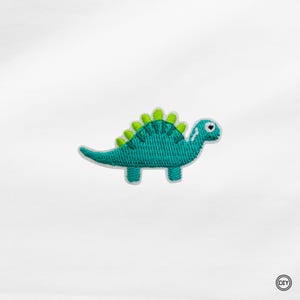 Könnte beinhalten: Gestickter Aufnäher mit einem türkisfarbenen Dinosaurier mit grünen Akzenten. Der Dinosaurier hat einen freundlichen Ausdruck und ist vor einem weißen Hintergrund platziert.