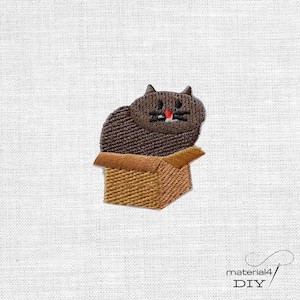 Könnte beinhalten: Ein gestickter Aufnäher mit einer dunkelbraunen Katze, die in einem beigefarbenen Pappkarton sitzt. Die Katze hat schwarze gestickte Augen, schwarze Schnurrhaare und eine kleine rote Nase. Der Karton hat eine schraffierte Textur.