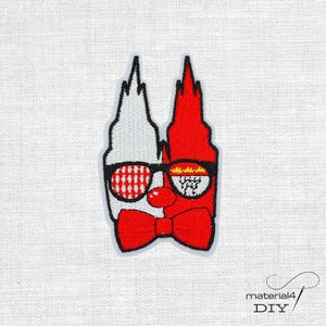 Könnte beinhalten: Ein gestickter Aufnäher mit zwei nebeneinander liegenden, spitzen Türmen, einer in Weiß und einer in Rot. Das Design zeigt eine schwarz umrandete Brille mit gemusterten Gläsern, eine kleine rote runde Nase und eine große rote Fliege.