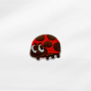 Könnte beinhalten: Ein kleiner, gestickter Aufnäher mit einem Cartoon-Marienkäfer. Der Marienkäfer hat einen roten Körper mit braunen Punkten, einen braunen Kopf und weiße Augen. Der Aufnäher ist zum Aufbügeln oder Aufnähen auf Stoff gedacht.