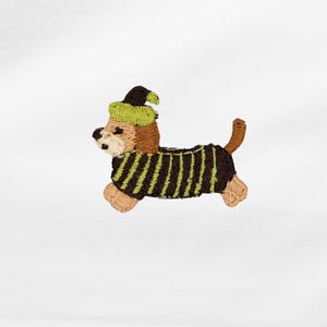 Könnte beinhalten: Gestickter Aufnäher eines Dackelhundes mit einem grünen und braunen Hexenhut und einem schwarz-grün gestreiften Pullover. Der Hund rennt. Der Aufnäher ist aus Faden gefertigt.