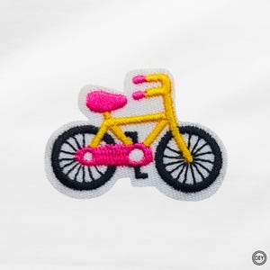 Könnte beinhalten: Gestickter Aufnäher mit einem gelben Fahrrad mit rosa Sattel und Akzenten. Das Fahrrad hat schwarze Räder mit weißen Speichen und ist vor einem weißen Hintergrund platziert. Ideal für DIY-Projekte.