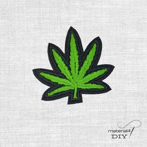 Könnte beinhalten: Gestickter Aufnäher mit einem grünen Cannabisblatt mit schwarzer Umrandung. Das Blattdesign ist auf einem weißen Stoffhintergrund zentriert. Der Aufnäher ist ein DIY-Bastelartikel.
