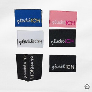 Könnte beinhalten: Eine Sammlung von sechs Stoffetiketten in verschiedenen Farben, darunter Blau, Rosa, Weiß und Schwarz. Jedes Etikett zeigt den gestickten Text "glücklICH" in verschiedenen Metallic-Fadenfarben wie Gold, Silber und Pink. Die Etiketten sind rechteckig und scheinen für Kleidung oder Bastelprojekte bestimmt zu sein.