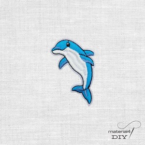 Könnte beinhalten: Gestickter Aufnäher mit einem springenden Delfin in Blau und Weiß. Der Delfin hat ein detailliertes Design mit weißem Bauch und blauem Umriss. Der Aufnäher ist wahrscheinlich für DIY-Projekte oder zur Individualisierung von Kleidung gedacht.