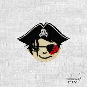Könnte beinhalten: Gestickter Aufnäher mit einem Cartoon-Piraten-Gesicht. Der Pirat trägt einen schwarzen Hut mit weißem Totenkopf und gekreuzten Knochen, eine rote Augenklappe und grüne Augen. Auf weißem Stoffhintergrund.