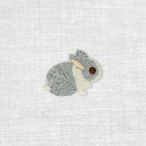 Könnte beinhalten: Gestickter Aufnäher eines kleinen Kaninchens. Das Kaninchen ist überwiegend grau mit einem weißen Streifen an der Seite und einem braunen Auge. Der Aufnäher befindet sich auf einem weißen Stoffhintergrund.