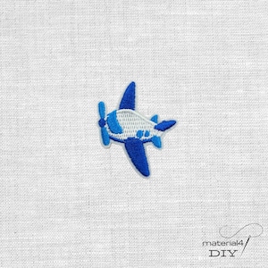 Könnte beinhalten: Ein gestickter Aufnäher in Form eines kleinen Flugzeugs. Das Flugzeug hat einen weißen Rumpf mit blauen Flügeln, einem blauen Heckbereich und einem blauen Propeller. Das Design ist in detaillierter Stickarbeit mit einem blauen Rand ausgeführt.