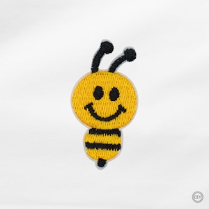Könnte beinhalten: Gestickter Aufnäher einer lächelnden Biene. Die Biene hat einen gelben Kopf und Körper, schwarze Streifen und schwarze Fühler. Das Gesicht hat zwei schwarze Augen und ein gebogenes Lächeln. Ideal für DIY-Projekte.