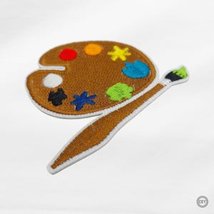 Könnte beinhalten: Gestickter Aufnäher mit einer braunen Künstlerpalette mit bunten Farbflecken und einem Pinsel. Die Palette hat rote, gelbe, blaue, grüne, schwarze und weiße Farbe. Der Aufnäher hat einen weißen Rand, für DIY-Projekte.