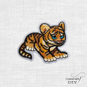 Könnte beinhalten: Gestickter Aufnäher eines Tigerbabys. Der Aufnäher hat orange und schwarze Streifen, blaue Augen und einen weißen Rand. Der Aufnäher befindet sich auf einem weißen Hintergrund. Der Text "material4 DIY" befindet sich in der unteren rechten Ecke.