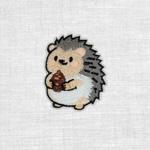 Könnte beinhalten: Gestickter Aufnäher mit einem niedlichen Igel, der einen braunen Tannenzapfen hält. Der Igel hat ein hellbraunes Gesicht und einen hellbraunen Bauch mit grauen und schwarzen Stacheln. Der Aufnäher ist weiß umrandet und befindet sich auf weißem Stoff.