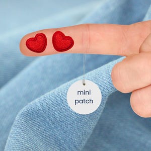 Könnte beinhalten: Zwei kleine rote herzförmige Aufnäher sind auf einem Finger abgebildet, mit einem weißen runden Anhänger mit der Aufschrift "mini patch", der darunter hängt. Die Aufnäher haben ein strukturiertes Aussehen und sind vor einem hellblauen Stoffhintergrund platziert.