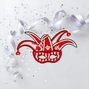 Könnte beinhalten: Ein rot-weißes Narrenmasken-Patch mit Harlekin-Design. Der Patch zeigt eine rote Maske mit weißen Akzenten und eine rot-weiße Narrenkappe. Die Maske ist von weißem Band und silbernen Konfetti umgeben.