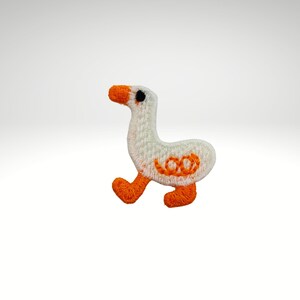 Könnte beinhalten: Gestickter Aufnäher einer weißen Gans mit orangefarbenem Schnabel, Füßen und dekorativen orangefarbenen Details. Die Gans wird beim Gehen mit einem schwarzen Auge dargestellt. Für Kleidung oder Accessoires.