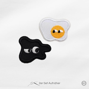 Könnte beinhalten: Zwei gestickte Aufnäher: einer schwarz, einer weiß mit gelber Mitte. Der schwarze Aufnäher hat weiße Augen, und der weiße Aufnäher ähnelt einem Spiegelei mit gelbem Eigelb und schwarzen Augen. Der Text unten lautet "2er Set Aufnäher".