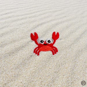 Könnte beinhalten: Ein roter, gestickter Krabbenaufnäher mit weißen Augen und einem lächelnden Mund. Die Krabbe hat große rote Scheren und befindet sich auf sandigem Untergrund. Der Aufnäher ist wahrscheinlich für Kleidung oder Accessoires.