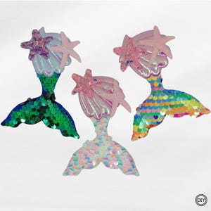 Könnte beinhalten: Drei Meerjungfrauen-Schwanz-Ornamente mit schillernden Muscheln und rosa Seestern-Akzenten. Ein Schwanz ist grün, ein anderer ist eine Mischung aus Pastellfarben, und der dritte ist eine Mischung aus Pastellfarben. Dekorative Ornamente.