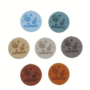 Könnte beinhalten: Sieben runde, handgefertigte Etiketten in verschiedenen Farben, darunter Blau, Grün, Beige, Grau, Braun und Orange. Jedes Etikett zeigt ein Bienen-Design mit Herz und dem Wort "HANDMADE". Die Etiketten bestehen aus einem strukturierten Material.