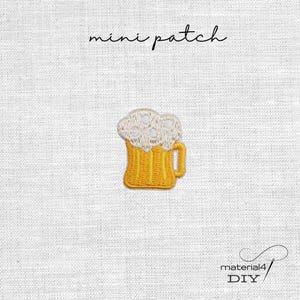 Könnte beinhalten: Ein gestickter Mini-Aufnäher in Form eines gelben Bierkrugs mit einer dicken weißen Schaumkrone. Der Text mini patch steht über dem Motiv und der Text material4 DIY befindet sich unten rechts auf einem weißen Stoffhintergrund.