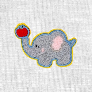 Könnte beinhalten: Ein grauer Elefantenaufnäher mit blauem Umriss und gelbem Rand, der einen roten Apfel hält. Der Elefant hat ein rosa Ohr und ein strukturiertes, flauschiges Aussehen. Der Aufnäher befindet sich auf einem weißen Stoffhintergrund.
