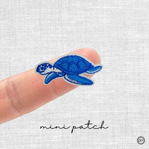 Könnte beinhalten: Ein kleiner, blau bestickter Aufnäher in Form einer Meeresschildkröte mit weißen Akzenten. Unter dem Aufnäher steht in Schreibschrift "mini patch". Der Aufnäher wird auf einem Finger gezeigt.