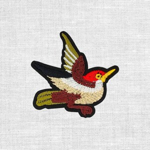 Könnte beinhalten: Gestickter Aufnäher mit einem bunten Vogel im Flug. Der Vogel hat einen roten Kopf, einen gelben Schnabel und Flügel mit grünen, weißen und braunen Details. Der Aufnäher ist schwarz umrandet und eignet sich für Kleidung oder Accessoires.