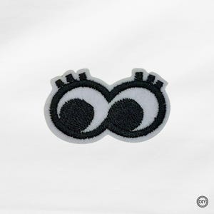 Könnte beinhalten: Gestickter Aufnäher mit einem Paar stilisierter Augen. Die Augen sind schwarz mit weißen Highlights und schwarzen Wimpern. Der Aufnäher hat einen weißen Rand und eignet sich für DIY-Projekte.