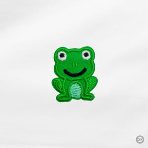 Könnte beinhalten: Ein grüner, gestickter Froschaufnäher mit einem lächelnden Gesicht und weißen Augen. Der Aufnäher ist ein DIY-Bastelartikel, ideal zum Hinzufügen zu Kleidung oder Accessoires. Der Frosch ist ein fröhliches Design.