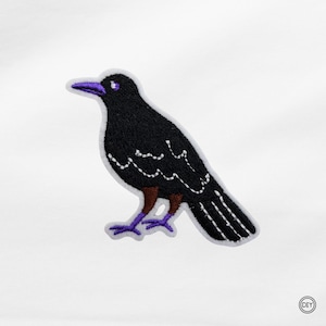 Könnte beinhalten: Gestickter Aufnäher mit einem schwarzen Raben, lila Schnabel, Füßen und Auge. Der Rabe hat weiße Nahtdetails und braune Akzente an den Beinen. Der Aufnäher hat einen weißen Rand und ist für DIY-Projekte geeignet.