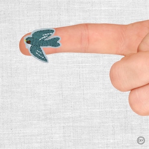 Könnte beinhalten: Ein teal-farbener Vogelaufnäher mit weißen Flügeldarstellungen wird auf einem Finger gezeigt. Der Aufnäher hat einen hellblauen Rand und einen braunen Schnabel. Der Hintergrund ist ein weißer Stoff.