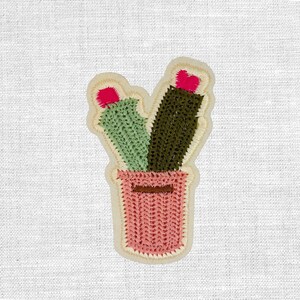Könnte beinhalten: Gestickter Aufnäher mit zwei Kakteen in einem rosa Topf. Ein Kaktus ist grün, der andere dunkelgrün, beide mit rosa Blütenakzenten. Der Aufnäher hat einen cremefarbenen Rand und ist vor einem weißen Stoffhintergrund platziert.
