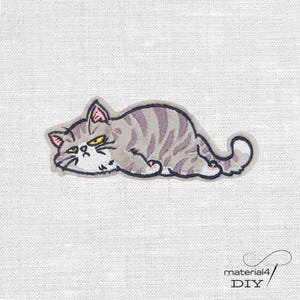 Könnte beinhalten: Gestickter Aufnäher mit einer Cartoon-Katze in entspannter Pose. Die Katze hat graues Fell mit lila Streifen, gelbe Augen und einen mürrischen Gesichtsausdruck. Der Aufnäher ist schwarz umrandet und hat einen weißen Hintergrund.