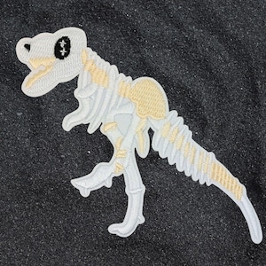 Könnte beinhalten: Gestickter Aufnäher eines Dinosaurierskeletts in Weiß und Beige. Der Dinosaurier hat schwarze Augen und ist vor einem dunklen Hintergrund platziert. Der Aufnäher ist detailliert mit Nähten.