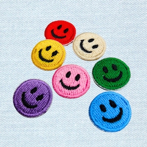 Könnte beinhalten: Eine Sammlung von sieben gestickten Smiley-Gesicht-Patches in verschiedenen Farben, darunter Rot, Gelb, Rosa, Lila, Grün, Beige und Blau. Jeder Patch hat ein schwarzes Smiley-Gesicht-Design. Die Patches sind kreisförmig und haben eine strukturierte Optik.