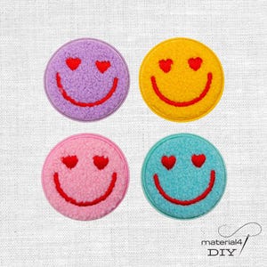 Könnte beinhalten: Vier kreisförmige Aufnäher mit einer flauschigen, strukturierten Oberfläche. Jeder Aufnäher zeigt ein Smiley-Gesicht mit zwei roten herzförmigen Augen und einem roten geschwungenen Mund. Die Aufnäher sind violett, gelb, hellrosa und hellblau.