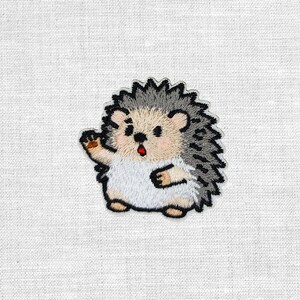 Könnte beinhalten: Gestickter Aufnäher mit einem winkenden Cartoon-Igel. Der Igel hat einen weißen Bauch, ein beiges Gesicht und graue Stacheln. Sein Mund ist geöffnet, und er hat eine kleine, braune Pfote zum Winken erhoben. Der Aufnäher ist schwarz umrandet.