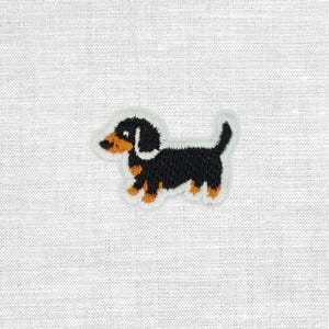 Könnte beinhalten: Ein gestickter Aufnäher, der einen kleinen Dackel mit schwarzem, hellbraunem und weißem Garn zeigt. Der Hund ist im Profil nach links blickend dargestellt, mit langem Körper, kurzen Beinen und erhobenem Schwanz, eingefasst von einem weißen Rand.