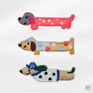 Könnte beinhalten: Drei gestickte Dackel-Patches. Der obere Patch ist rosa mit weißen Punkten, der mittlere ist beige mit blauen und roten Punkten und der untere ist beige mit einem weiß-schwarz gefleckten Mantel. Jedes Patch hat ein anderes Design.