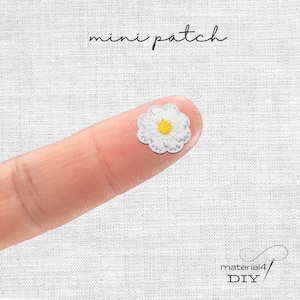 Könnte beinhalten: Ein kleiner, gestickter Aufnäher in Form einer Gänseblume mit weißen Blütenblättern und einem gelben Zentrum. Der Aufnäher wird auf einem Finger gehalten, darüber steht "mini patch". Der Hintergrund ist ein strukturiertes weißes Gewebe.