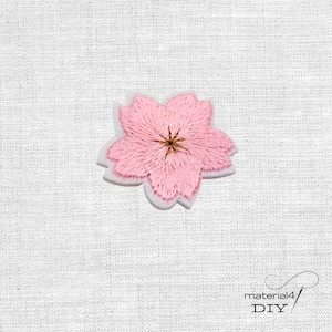 Könnte beinhalten: Ein rosa gestickter Blumenaufnäher mit weißem Rand. Die Blume hat fünf Blütenblätter und eine braune Mitte. Der Aufnäher befindet sich auf einem weißen, strukturierten Hintergrund. Der Text "material4 DIY" befindet sich unten rechts.