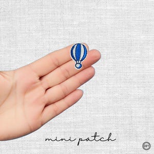 Könnte beinhalten: Ein kleiner, gestickter Aufnäher mit einem blau-weißen Heißluftballon-Design. Der Aufnäher wird in einer Hand vor einem weißen Stoffhintergrund gehalten. Die Worte "mini patch" sind darunter in Schreibschrift geschrieben.