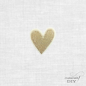 Könnte beinhalten: Ein kleiner herzförmiger Aufnäher mit metallisch goldener Stickerei und einem dichten, strukturierten Stichmuster. Das Herz hat einen hellen Rand und ist auf einem schlichten, cremeweißen Webstoff mit sichtbaren Textilfasern zentriert.