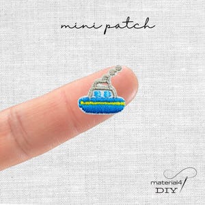 Könnte beinhalten: Ein kleiner gestickter Aufnäher, der ein blaues Boot mit einem gelben Streifen und einer grauen Rauchfahne zeigt. Der Text 'mini patch' steht über dem Boot und der Text 'material4 DIY' befindet sich in der unteren rechten Bildecke.