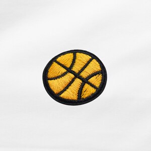 Könnte beinhalten: Runder, gestickter Aufnäher mit einem Basketball-Design. Der Aufnäher hat einen schwarzen Rand. Der Basketball ist gelb mit schwarzen Linien, die die Felder des Balls darstellen. Geeignet für Kleidung oder Accessoires.