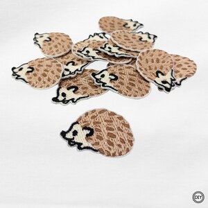 Könnte beinhalten: Eine Ansammlung von gestickten Igel-Patches. Jeder Patch hat einen braunen Körper mit einem strukturierten Muster, ein weißes Gesicht und schwarze Augen. Die Patches sind auf weißem Hintergrund verstreut.