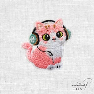 Könnte beinhalten: Ein gestickter Aufnäher, der eine rosa-weiße Katze mit türkisfarbenen und schwarzen Kopfhörern zeigt. Die Katze hat große grüne Augen, eine kleine rosa Nase und ein schwarzes gesticktes Kabel, das vom Kopfhörer zum Körper führt, auf weißem Stoff.