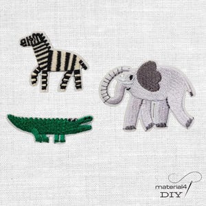 Könnte beinhalten: Drei bestickte Stoffaufnäher mit Tiermotiven: ein schwarz-weiß gestreiftes Zebra, ein grauer Elefant mit einem dunkelgrauen Ohr und ein grünes Krokodil. Der Text material4 DIY ist in der unteren rechten Ecke des Bildes zu sehen.