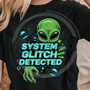 Könnte beinhalten: Schwarzes T-Shirt mit einem grünen Alien-Design, großen schwarzen Augen und grünen Händen. Der Text "SYSTEM GLITCH DETECTED" ist türkis. Zwei UFOs sind ebenfalls im Design. Das Shirt ist aus weichem Material.